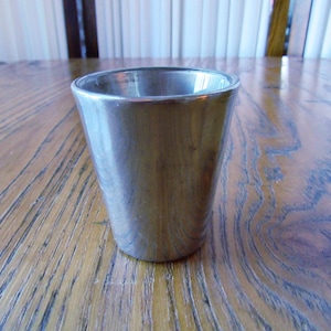 May include: A silver-coloured, tapered drinking glass. The glass has a wide mouth and a narrow base. The glass is on a wooden table with a natural wood grain pattern. The background is blurred.