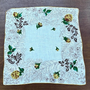 May include: Square white handkerchief with a floral design. The handkerchief features yellow roses, green leaves, and brown and white floral accents. The edges are trimmed with a gold border.
