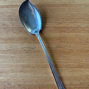 Peut inclure: Une cuillère de service argentée avec une cuillère en forme de feuille et un long manche fin. La cuillère repose sur une surface en bois, mettant en valeur sa finition polie et l'extrémité décorative de la poignée.