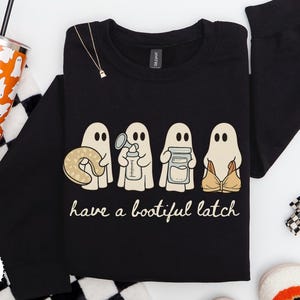 May include: A black long-sleeve sweatshirt featuring a graphic of four ghosts. The ghosts are holding a nursing pillow, a breast pump, a milk storage jar, and a nursing bra. Below the ghosts, the text reads, have a bootiful latch.