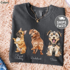 May include: Dark grey Comfort Colors t-shirt with watercolour illustrations of a French Bulldog, Dachshund, and Terrier. Breed names are below each dog. A gold necklace is visible.