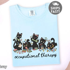 May include: A light blue t-shirt featuring a graphic of five black cats engaged in various craft activities. The cats are surrounded by small items like yarn, beads, and scissors. Below the cats, the text reads occupational therapy.
