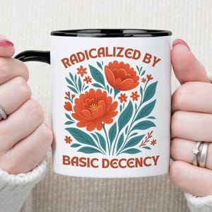 May include: White ceramic mug with a black interior and handle, featuring the text "RADICALIZED BY BASIC DECENCY" in orange. The mug is decorated with orange flowers and teal leaves.