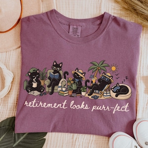 May include: A berry-coloured t-shirt featuring a graphic of five black cats engaged in various leisure activities, including reading, drinking, and relaxing. The text below the cats reads, "retirement looks purr-fect" in a white cursive script font.