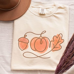 May include: Ivory-colored t-shirt featuring a line drawing of an acorn, pumpkin, and leaf in shades of orange and brown. A brown hat is visible in the upper left corner. The shirt has a Comfort Colors tag.