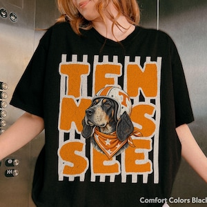 May include: A black t-shirt featuring a graphic of a hound dog wearing a white helmet with an orange stripe and an orange bandana with white stars. The word TEN SEE is printed in large, distressed orange block letters over vertical white stripes.