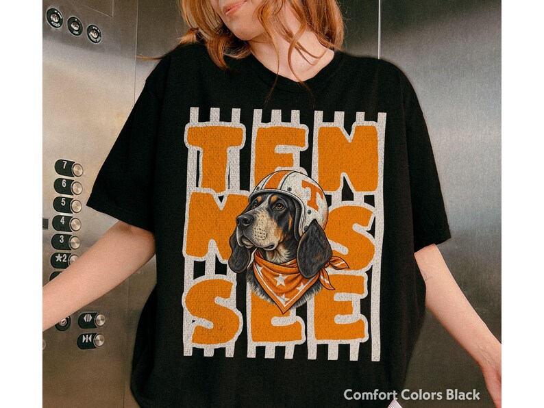 May include: A black t-shirt featuring a graphic of a hound dog wearing a white helmet with an orange stripe and an orange bandana with white stars. The word TEN SEE is printed in large, distressed orange block letters over vertical white stripes.