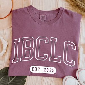 May include: Berry-colored t-shirt with the white text "IBCLC" in a collegiate font and "EST. 2025" below. The shirt is displayed on a neutral-toned surface with a hat, scrunchie, and shoes.