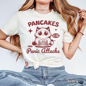 May include: A white t-shirt with a red graphic of a cartoon cat with a stack of pancakes and the text "Pancakes and Panic Attacks" with the words "Buy One Get One" in a smaller font above the cat.