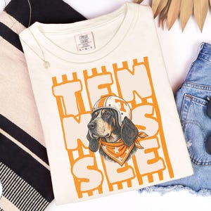 May include: An ivory short-sleeve t-shirt featuring a graphic of a hound dog wearing a white and orange helmet and an orange bandana with white stars. The text TEN SEE is printed in large orange block letters behind the dog illustration.