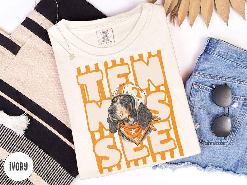 May include: An ivory short-sleeve t-shirt featuring a graphic of a hound dog wearing a white and orange helmet and an orange bandana with white stars. The text TEN SEE is printed in large orange block letters behind the dog illustration.