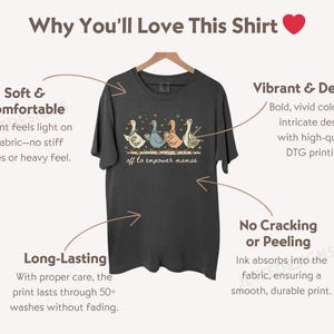 May include: Dark gray t-shirt with a whimsical design of three geese holding musical instruments, with the text "off to empower mamas." The shirt is displayed on a wooden hanger.