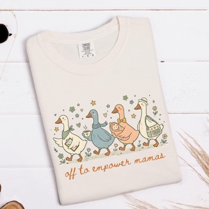 May include: Ivory-colored t-shirt featuring a whimsical illustration of geese walking in a line, each with unique accessories. The text "off to empower mamas" is written in a cursive font. The shirt is a Comfort Colors brand.