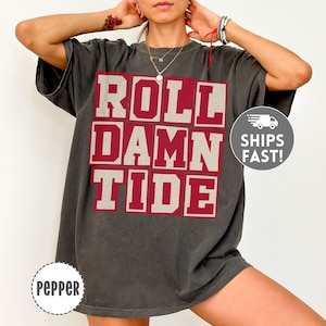 May include: A charcoal grey short-sleeve t-shirt featuring the text ROLL DAMN TIDE in large, block-style maroon letters with a light grey outline. The shirt has a relaxed, oversized fit and a crew neckline.