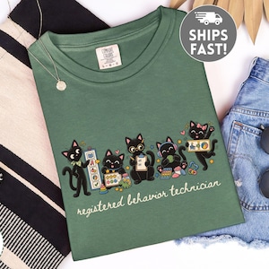 May include: A moss green crew neck t-shirt featuring a graphic of five black cats engaged in various activities, with the text "registered behavior technician" printed in white below the cats. The shirt has a Comfort Colors brand label at the neck.