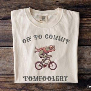 May include: Ivory-colored t-shirt with the text "OFF TO COMMIT TOMFOOLERY." The shirt features a cartoon frog riding a bicycle, wearing a scarf. The shirt is folded on a wooden surface.