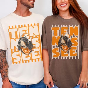 May include: An ivory t-shirt and an espresso t-shirt, each featuring a graphic of a hound dog wearing a football helmet and a bandana. The text "TEN SEE" is printed in large orange letters behind the dog, set against a background of vertical stripes.