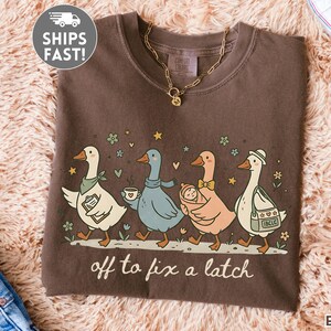 May include: Espresso-colored t-shirt featuring a whimsical illustration of five geese walking in a row. Each goose is uniquely accessorized, carrying items like a clipboard, coffee, a baby, and a bag. The text "off to fix a latch" is printed below the geese.