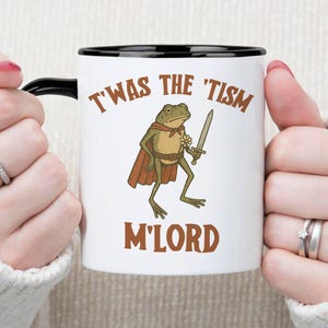 May include: White ceramic mug with a black interior and handle. The mug features a cartoon frog wearing a cape and holding a sword, with the text "T'WAS THE 'TISM M'LORD" in brown lettering.