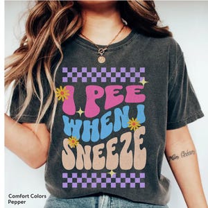 May include: Pepper-colored t-shirt with the text "I PEE WHEN I SNEEZE" in a retro font. The design includes a checkerboard pattern, flowers, and stars. The shirt is a comfortable, casual wear.