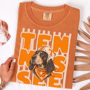 May include: A burnt orange t-shirt featuring a graphic of a hound dog wearing a white helmet with an orange T and an orange bandana with white stars. The text TEN SEE is printed in large orange block letters behind the dog illustration.