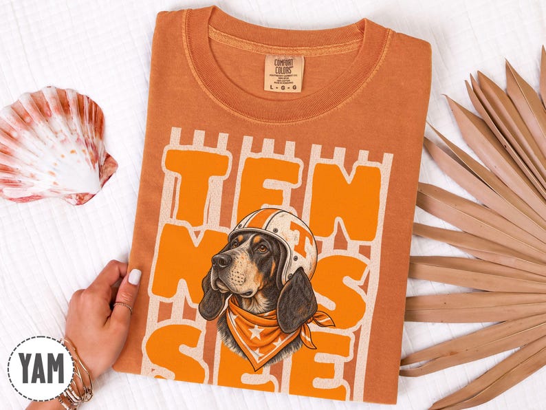 May include: A burnt orange t-shirt featuring a graphic of a hound dog wearing a white helmet with an orange T and an orange bandana with white stars. The text TEN SEE is printed in large orange block letters behind the dog illustration.