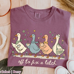 May include: Berry-colored t-shirt with a whimsical illustration of four geese walking in a row. The geese are carrying various items, including a baby, coffee, and a briefcase. The text "off to fix a latch" is printed below the geese.