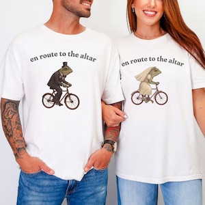 May include: White t-shirts with a whimsical design featuring frogs riding bicycles. One frog wears a suit and top hat, the other a wedding dress and veil. The text "en route to the altar" is printed above the illustrations.