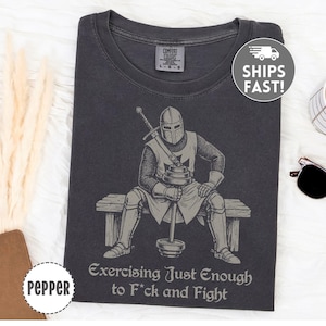 May include: Dark gray t-shirt featuring a knight lifting weights, with the text "Exercising Just Enough to F*ck and Fight." The shirt has a vintage look and a crew neck. The shirt is a Comfort Colors brand.