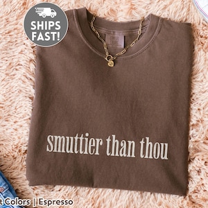 Puede incluir: Camiseta marr&oacute;n con la frase "smuttier than thou" en letras blancas. Un collar de cadena dorada con un colgante descansa sobre la camiseta. La camiseta est&aacute; sobre una superficie suave y marr&oacute;n claro.