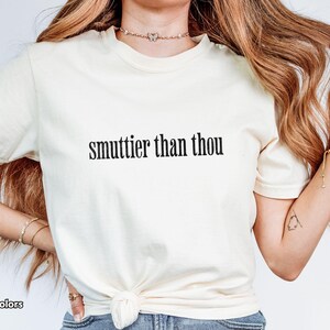 Puede incluir: Camiseta de color marfil con la frase "smuttier than thou" impresa en negro. La camiseta es de la marca Comfort Colors. La modelo lleva la camiseta y tiene el pelo largo y ondulado.