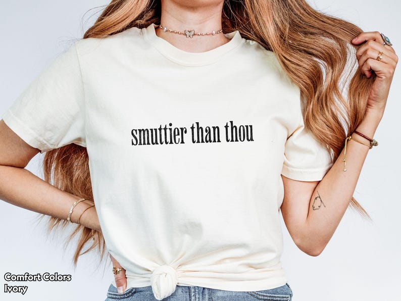 Puede incluir: Camiseta de color marfil con la frase "smuttier than thou" impresa en negro. La camiseta es de la marca Comfort Colors. La modelo lleva la camiseta y tiene el pelo largo y ondulado.