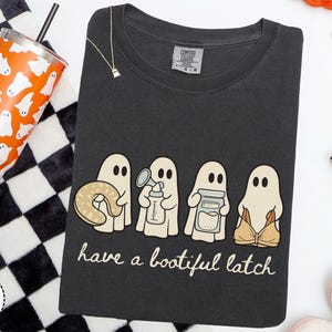 May include: A charcoal gray t-shirt featuring a graphic of four ghosts holding a nursing pillow, a breast pump, a storage jar, and a nursing bra. The text below the ghosts reads have a bootiful latch in a white script font. The shirt has a tag.