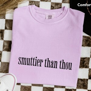 Puede incluir: Una camiseta Comfort Colors de color lila claro con la frase "smuttier than thou" en negro. La camiseta se muestra sobre una superficie a cuadros blancos y negros, con zapatillas blancas y bolas de discoteca.