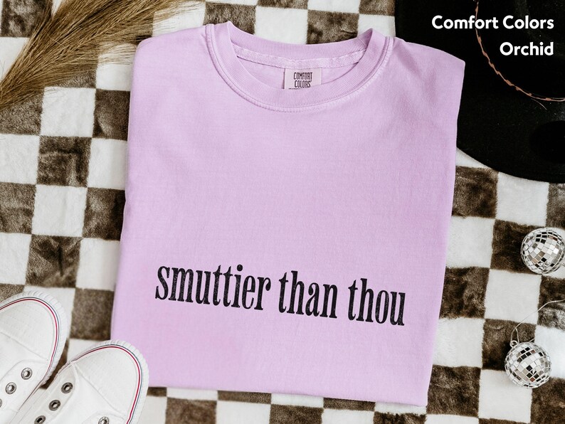 Puede incluir: Una camiseta Comfort Colors de color lila claro con la frase "smuttier than thou" en negro. La camiseta se muestra sobre una superficie a cuadros blancos y negros, con zapatillas blancas y bolas de discoteca.