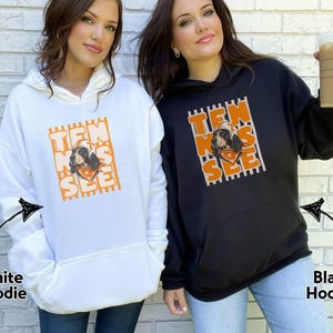 May include: A white hoodie and a black hoodie, each featuring a graphic print of a dog wearing a hat and bandana. The text "TEN NES SEE" is arranged vertically in orange block letters over a background of vertical stripes on both garments.