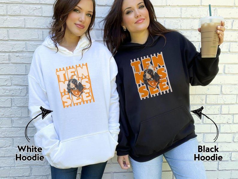 May include: A white hoodie and a black hoodie, each featuring a graphic print of a dog wearing a hat and bandana. The text "TEN NES SEE" is arranged vertically in orange block letters over a background of vertical stripes on both garments.