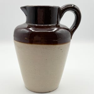 May include: A vintage stoneware pitcher with a dark brown top and handle, and a light beige body. The pitcher has a classic, rounded shape with a spout for pouring. The handle is arched and sturdy. A decorative piece for the kitchen.