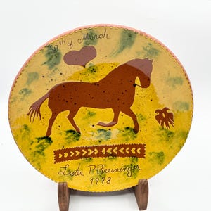 May include: A round ceramic plate with a brown horse design, dated 1998. The plate has a yellow background with green and brown accents, and the text "30th of March" is written on the top. The artist's name, Lester P. Breininger, is also visible.