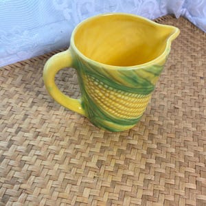May include: A ceramic creamer pitcher featuring a molded corn cob design with a yellow interior and exterior surface. The exterior displays textured yellow corn kernels surrounded by green husks, complete with a curved handle and a pouring spout.