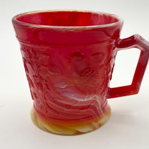 May include: A red glass mug with a decorative bird and floral pattern. The mug has a square handle and a flared base. The glass has a swirled, iridescent effect with shades of red, orange, and white.