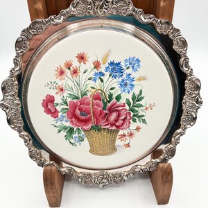 May include: A silver-plated serving tray with a floral design. The tray features a white center with a painted image of a basket filled with red peonies, blue cornflowers, and other colorful flowers. The tray has a decorative, ornate edge.