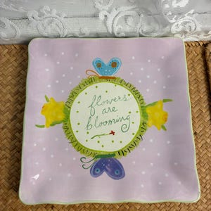 May include: A square ceramic plate with a light purple background featuring white polka dots. The center displays a circular design with the text "flowers are blooming" in green script, surrounded by two yellow flowers and two butterflies in blue and purple.