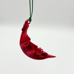 May include: A glossy, deep red crescent moon ornament with a sculpted face featuring closed eyes and a nose. The ornament is suspended by a thin, dark green string attached to a small loop at the top of the moon's curve, creating a hanging decoration.