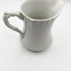 May include: A white ceramic pitcher with a unique, textured handle. The pitcher has a curved spout and a slightly irregular rim. The overall design is simple and elegant, suitable for serving beverages or as a decorative piece.