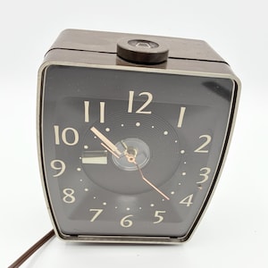May include: A vintage brown alarm clock with a black face and silver trim. The clock features white numerals and hands, with a small button on top. The clock is plugged in with a brown cord.