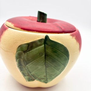 May include: A ceramic apple-shaped lidded jar with a cream-colored base featuring a fine crackle glaze. The lid is painted in a deep red hue with a dark green stem. A prominent, textured green leaf is molded onto the front of the apple body.