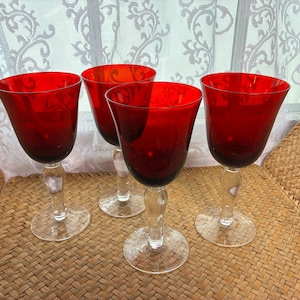 May include: A set of four wine glasses featuring deep red glass bowls and clear glass stems with circular bases. The bowls have a smooth, rounded shape that tapers toward the base, while the stems are thick and transparent, providing a contrast.