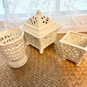 May include: A set of three off-white ceramic pieces featuring intricate cut-out lattice patterns. The collection includes a cylindrical footed vessel, a square lidded box with a pointed finial, and a square open-top container with four small feet.