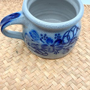 May include: A light gray ceramic mug with a speckled texture and a rounded body. The exterior features a raised, embossed floral pattern in deep blue glaze. A single handle is attached to the side, also accented with a deep blue glaze finish.